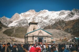 A crowd of pilgrims at Kedarnath Temple with snowcapped Himalayan peaks in the background.