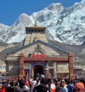 Crowds gather at the iconic Kedarnath Temple against a majestic Himalayan backdrop in daylight.