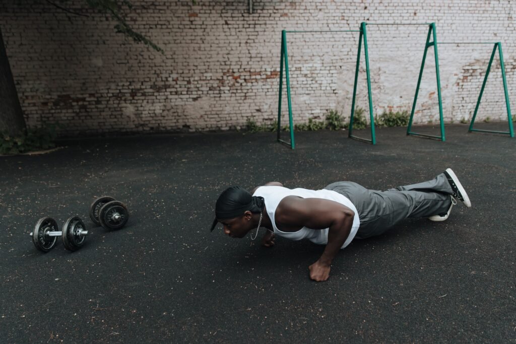 A man in sports attire doing push-ups on an outdoor asphalt surface beside dumbbells.