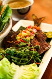 Vibrant dish of spicy grilled fish topped with vegetables served alongside fresh greens and soup.